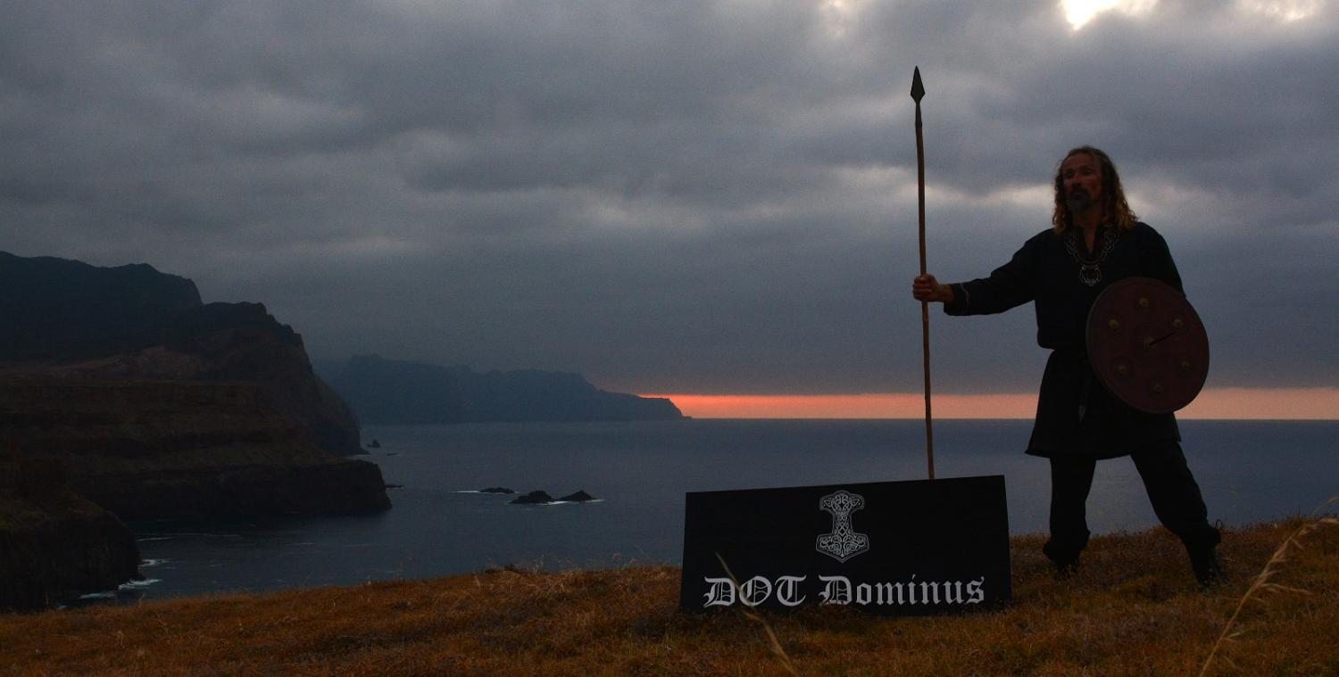 DOT Dominus on the Madeira cliffs at dusk, spear and round shield, Mjölnir banner at his feet